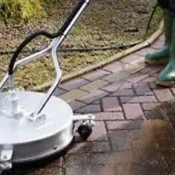 Czyszczenie kostki brukowej za pomocą myjki ciśnieniowej z okrągłą dyszą, widoczne zielone kalosze operatora i mokra, umyta część powierzchni.