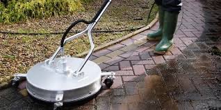 Czyszczenie kostki brukowej za pomocą myjki ciśnieniowej z okrągłą dyszą, widoczne zielone kalosze operatora i mokra, umyta część powierzchni.