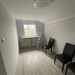 Graf Transport-Remonty - Pokój po malowaniu na szaro, widoczna drabina, dwa krzesła, okno z roletą i grzejnik