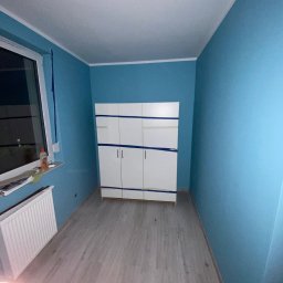 Graf Transport-Remonty - Pokój po malowaniu na niebiesko, z zabezpieczoną taśmą białą szafą i grzejnikiem pod oknem.