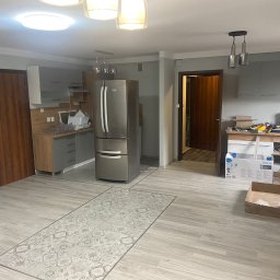 Graf Transport-Remonty - Wnętrze kuchni w trakcie odświeżania, widoczne szare szafki, srebrna lodówka, panele podłogowe z wzorem dywanu, otwarte drzwi do innego pomieszczenia i narzędzia malarskie na stole.