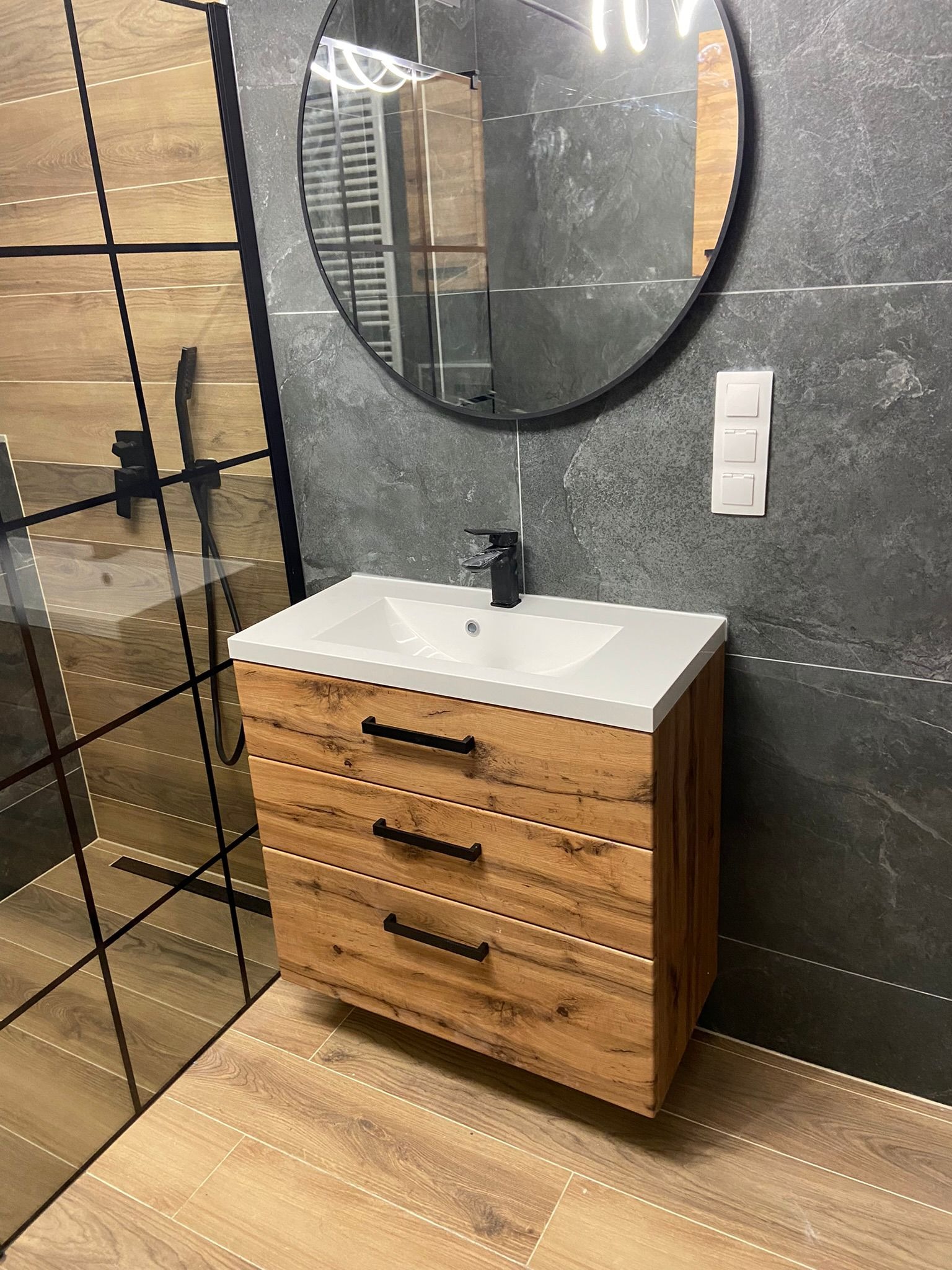 Nowoczesna łazienka z drewnianą szafką pod umywalką, czarną armaturą i szarą ścianą z efektem kamienia, oraz kabiną prysznicową z drewnopodobnymi płytkami.