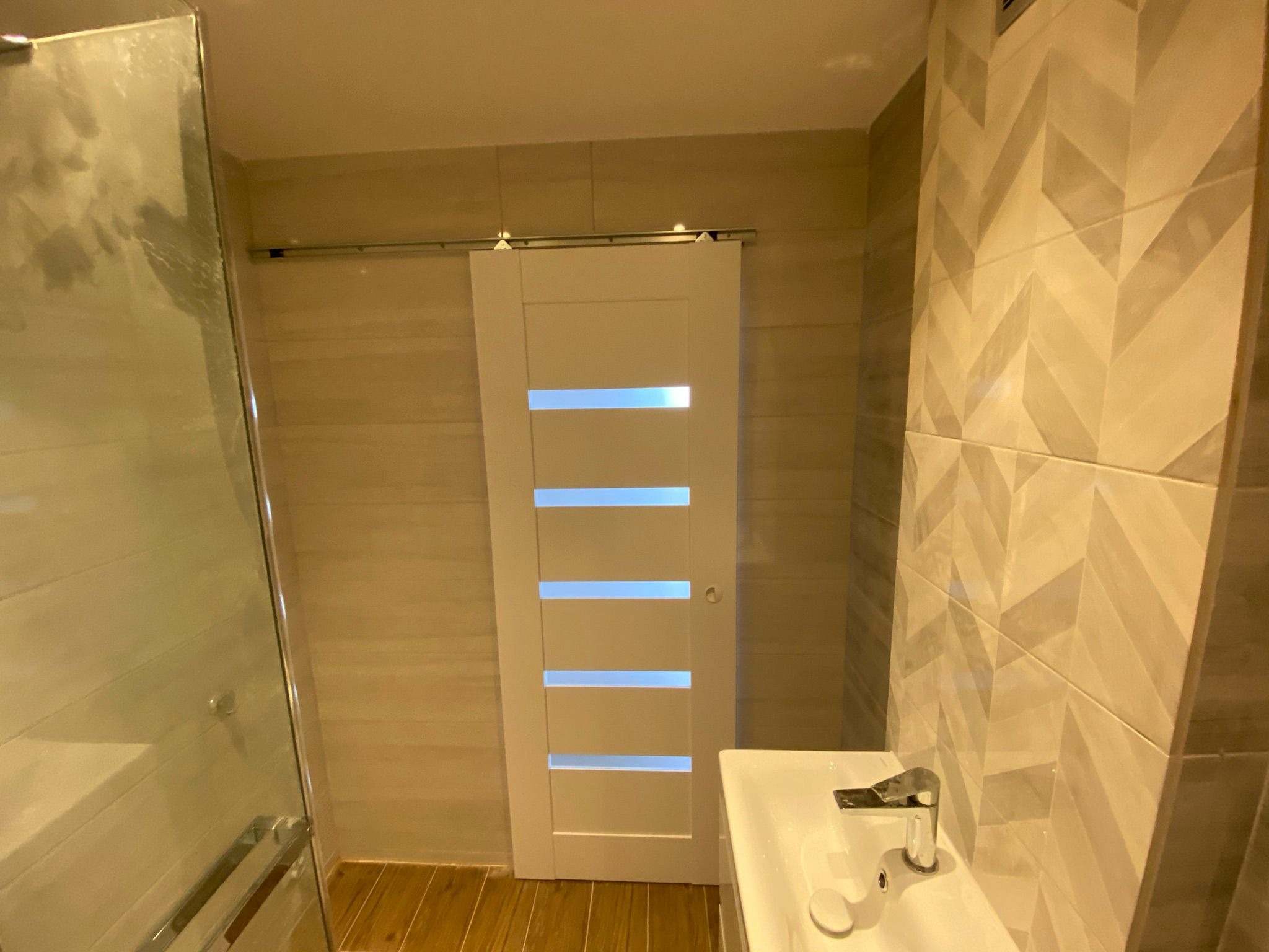 Nowoczesna łazienka z przesuwnymi drzwiami z czterema poziomymi przeszkleniami, umywalką z chromowaną baterią i ścianą wyłożoną płytkami w jodełkę.