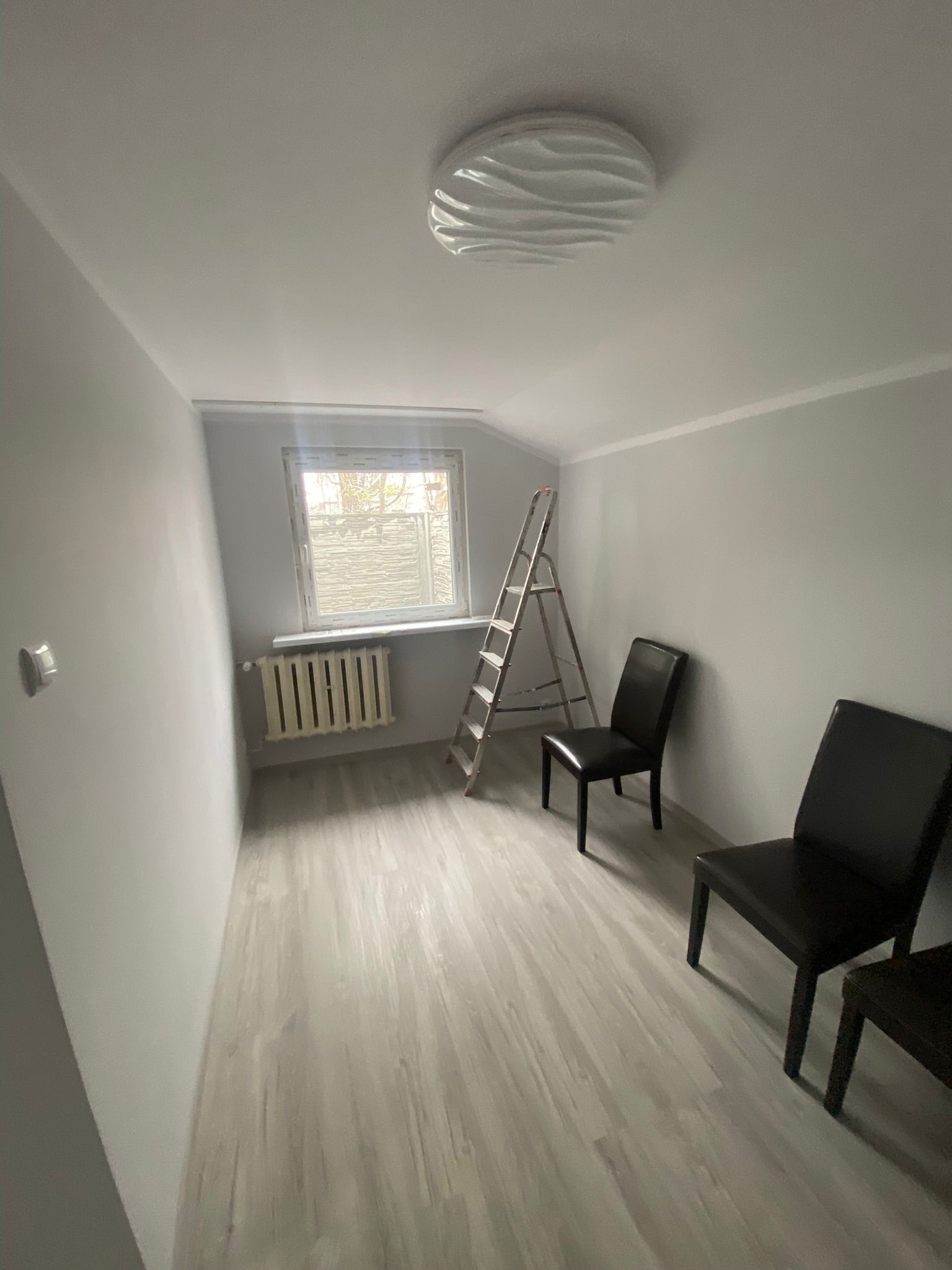 Pokój po malowaniu na szaro, widoczna drabina, dwa krzesła, okno z roletą i grzejnik