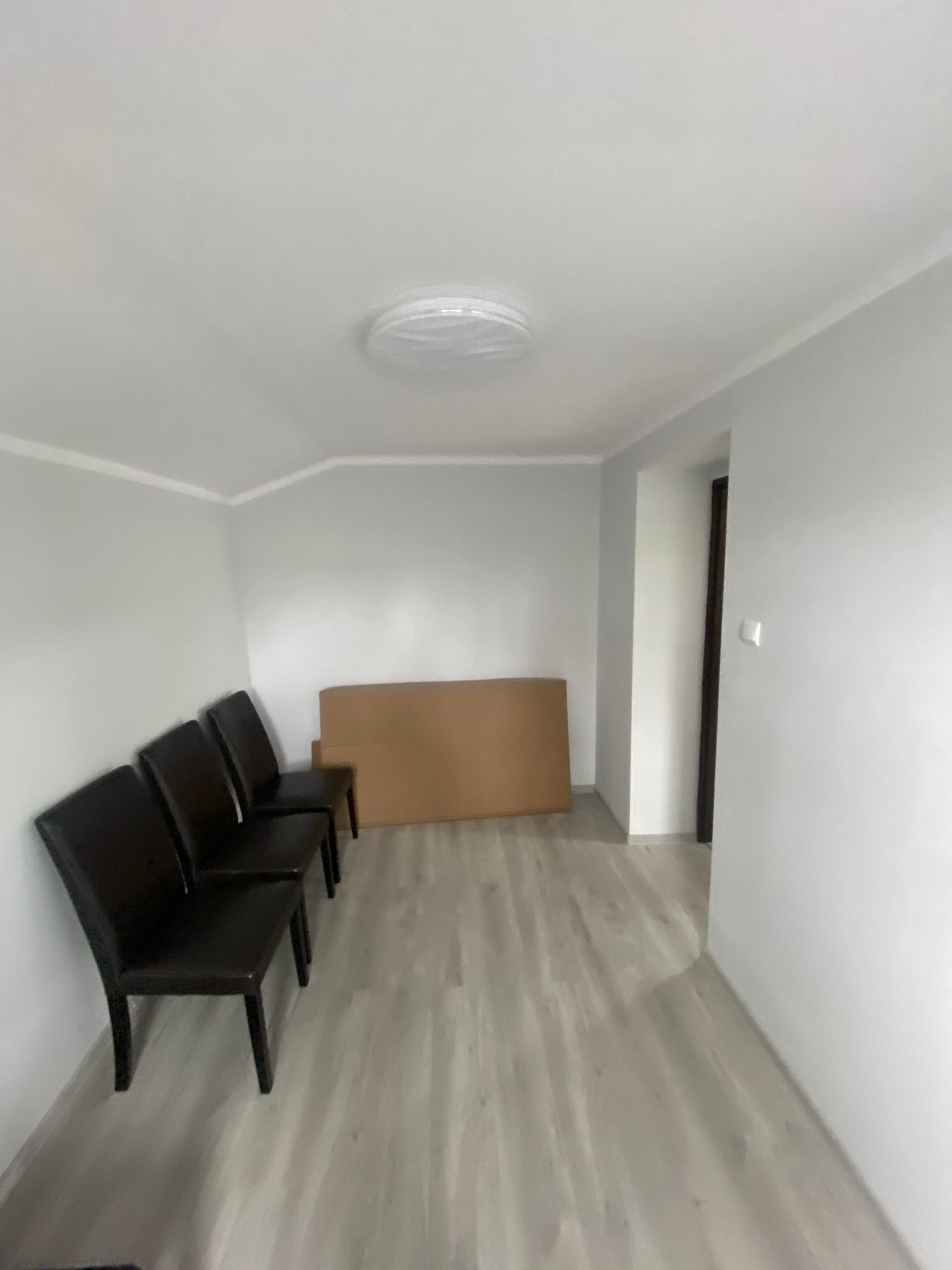 Puste, jasne pomieszczenie z szarymi panelami podłogowymi, czterema ciemnymi krzesłami ustawionymi pod ścianą i kartonami w tle, oświetlone okrągłą lampą sufitową.