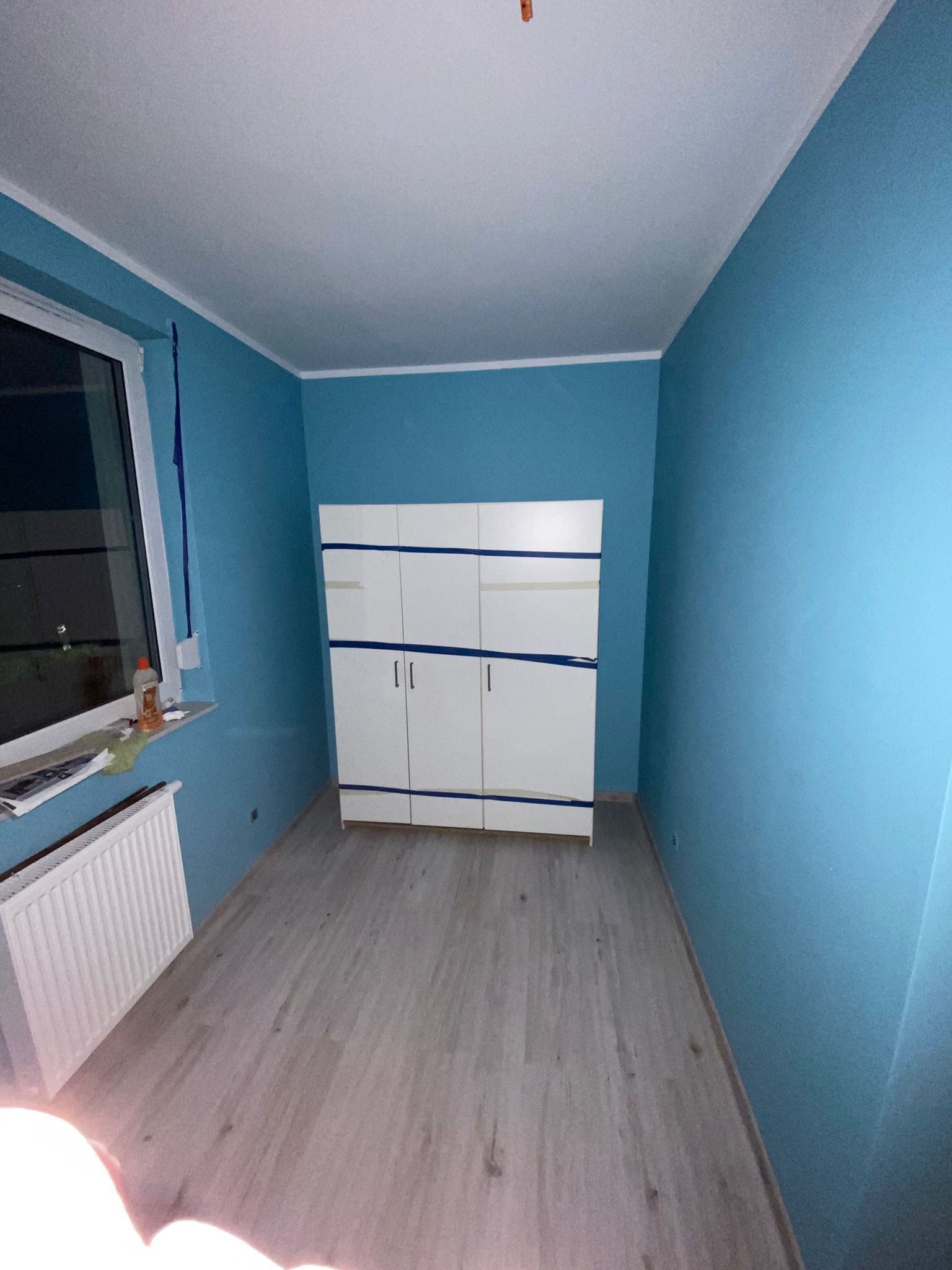 Pokój po malowaniu na niebiesko, z zabezpieczoną taśmą białą szafą i grzejnikiem pod oknem.
