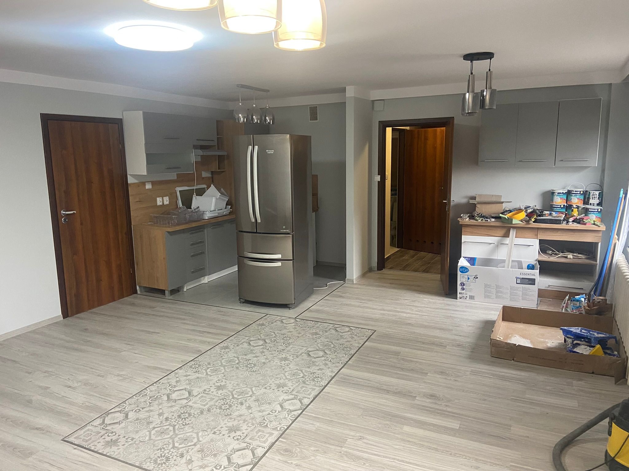 Wnętrze kuchni w trakcie odświeżania, widoczne szare szafki, srebrna lodówka, panele podłogowe z wzorem dywanu, otwarte drzwi do innego pomieszczenia i narzędzia malarskie na stole.