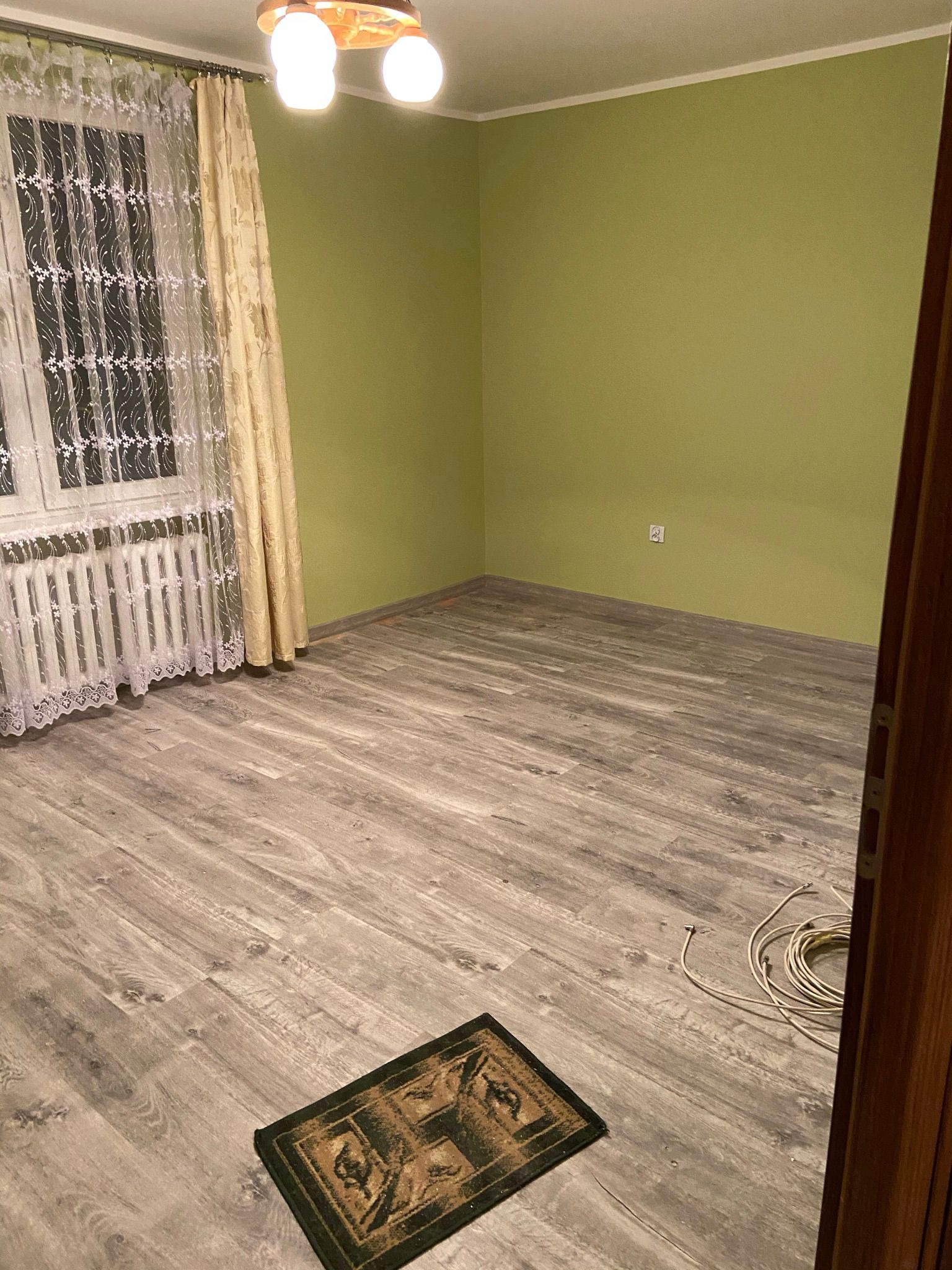 Pusty pokój z zielonymi ścianami, panelami podłogowymi w odcieniach szarości, grzejnikiem pod oknem z firankami i dywanikiem przy drzwiach, widoczne kable na podłodze.