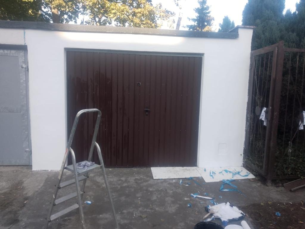 Świeżo pomalowany garaż z brązową bramą segmentową, widoczna drabina aluminiowa i resztki farby na betonowej posadzce.
