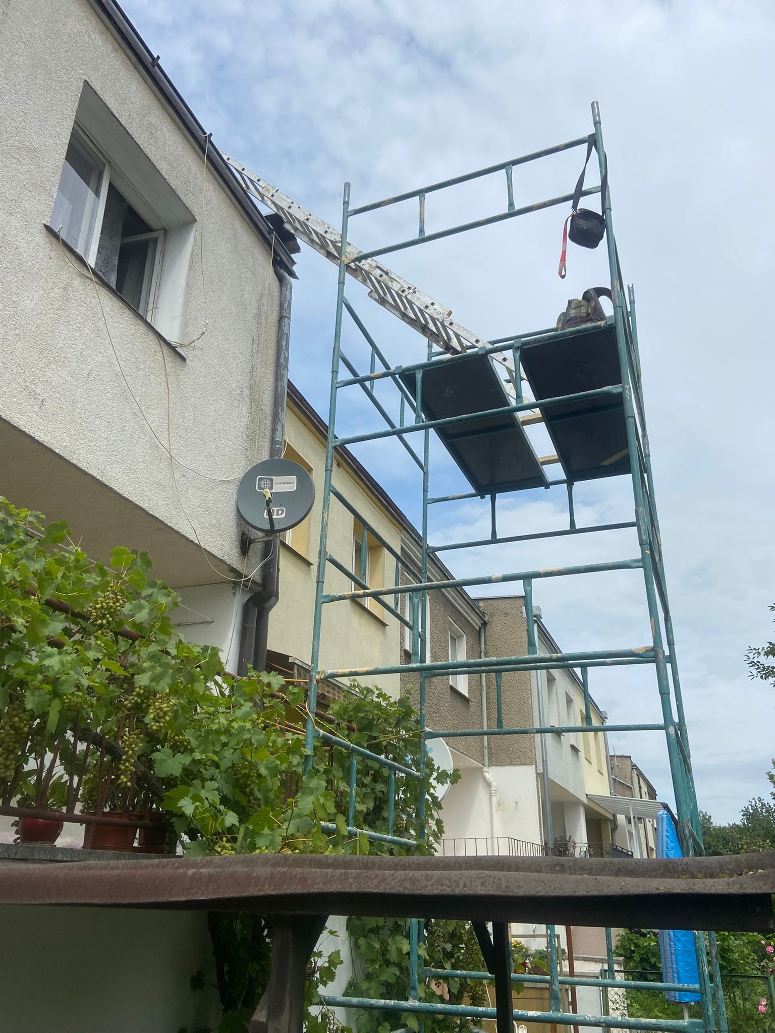 Rusztowanie przy elewacji budynku wielorodzinnego, widoczna antena satelitarna, balkon z pnączami winogron, niebieskie niebo.