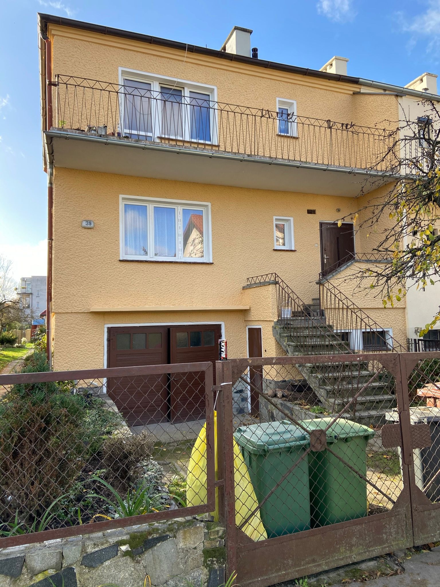 Żółta kamienica z balkonem, schodami zewnętrznymi i garażem, widoczna zza metalowej siatki ogrodzeniowej i dwóch zielonych pojemników na śmieci.