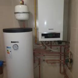 Biały piec gazowy marki Buderus zamontowany na ścianie z beżowych płytek, połączony miedzianymi rurami z białym zasobnikiem ciepłej wody Kospel, widoczny manometr i wyświetlacz pieca.