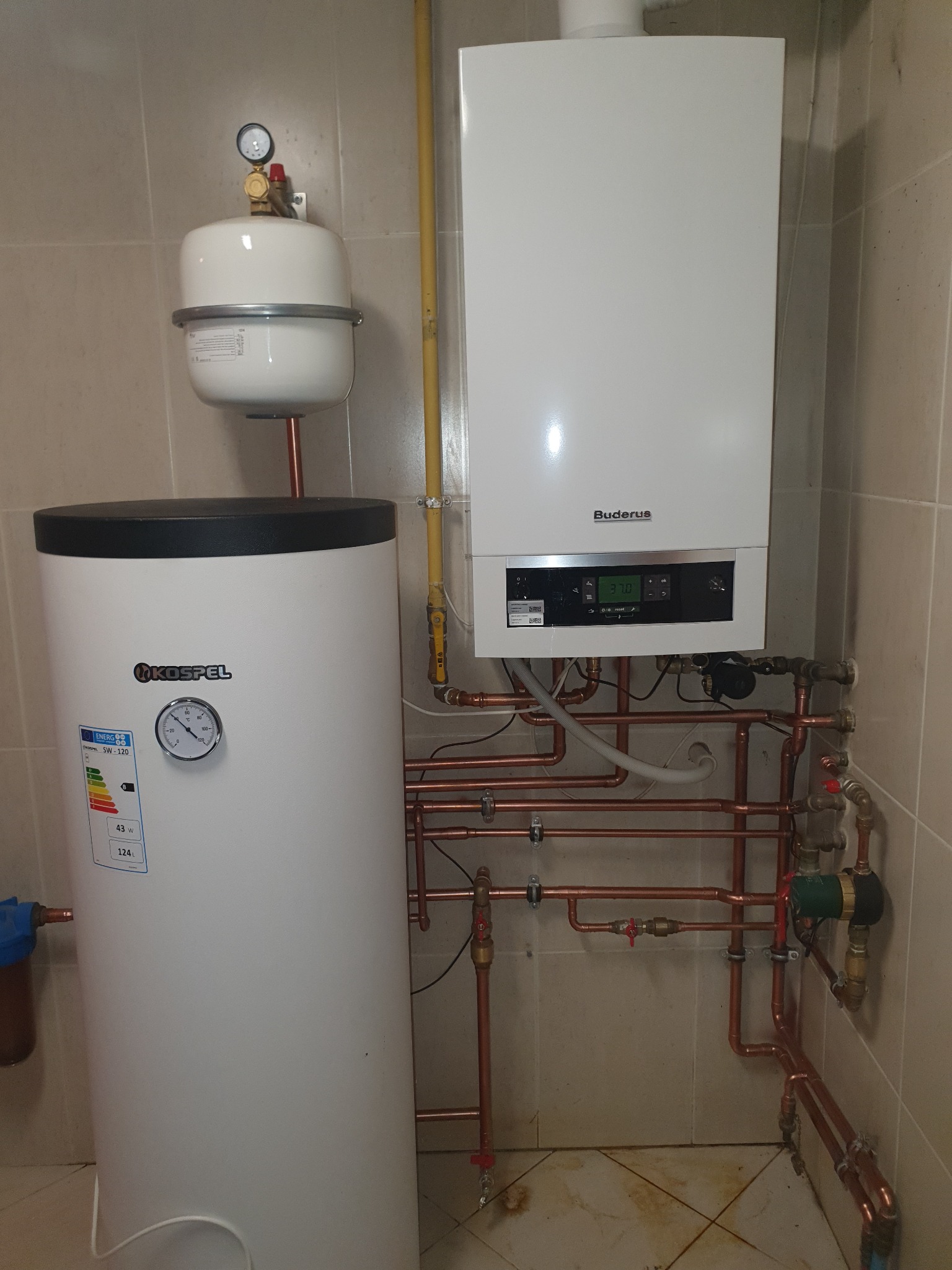 Biały piec gazowy marki Buderus zamontowany na ścianie z beżowych płytek, połączony miedzianymi rurami z białym zasobnikiem ciepłej wody Kospel, widoczny manometr i wyświetlacz pieca.