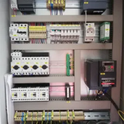 Rozdzielnica elektryczna z widocznymi bezpiecznikami, przekaźnikami, sterownikami Siemens, wyłącznikami Eaton, gniazdem elektrycznym i przewodami oznaczonymi numerami, zamontowana w metalowej...