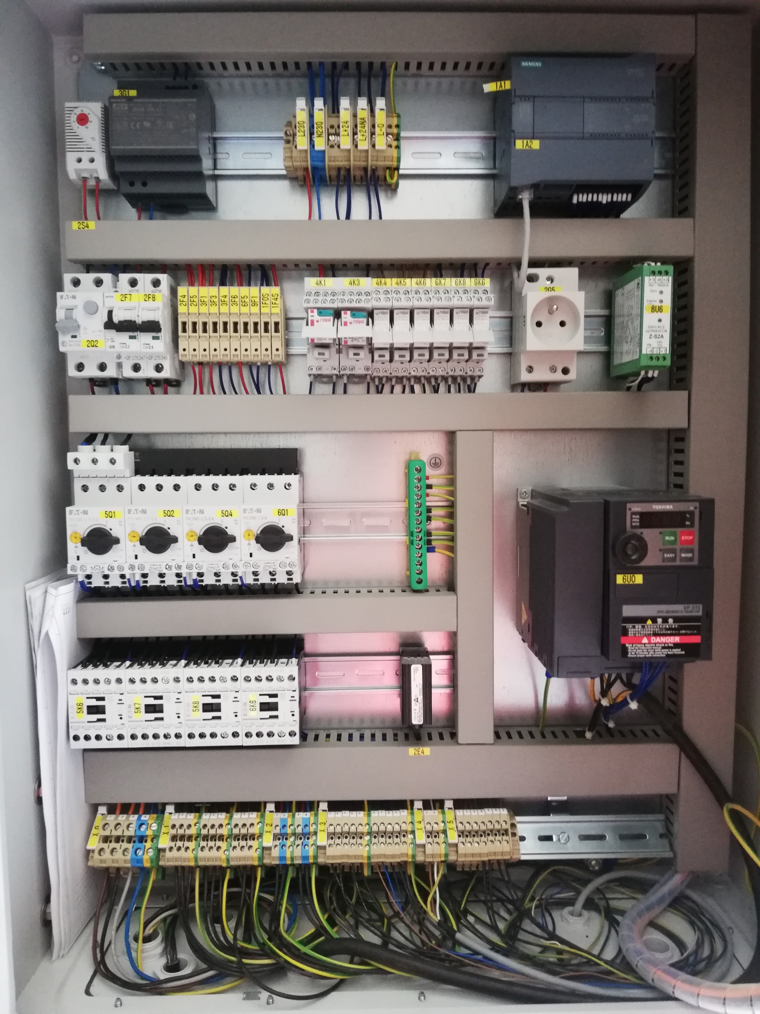 Rozdzielnica elektryczna z widocznymi bezpiecznikami, przekaźnikami, sterownikami Siemens, wyłącznikami Eaton, gniazdem elektrycznym i przewodami oznaczonymi numerami, zamontowana w metalowej...