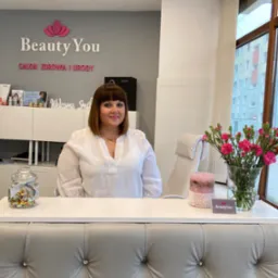 Recepcjonistka w salonie kosmetycznym Beauty You, za ladą z dekoracyjnym obiciem, w tle logo i wystrój wnętrza.