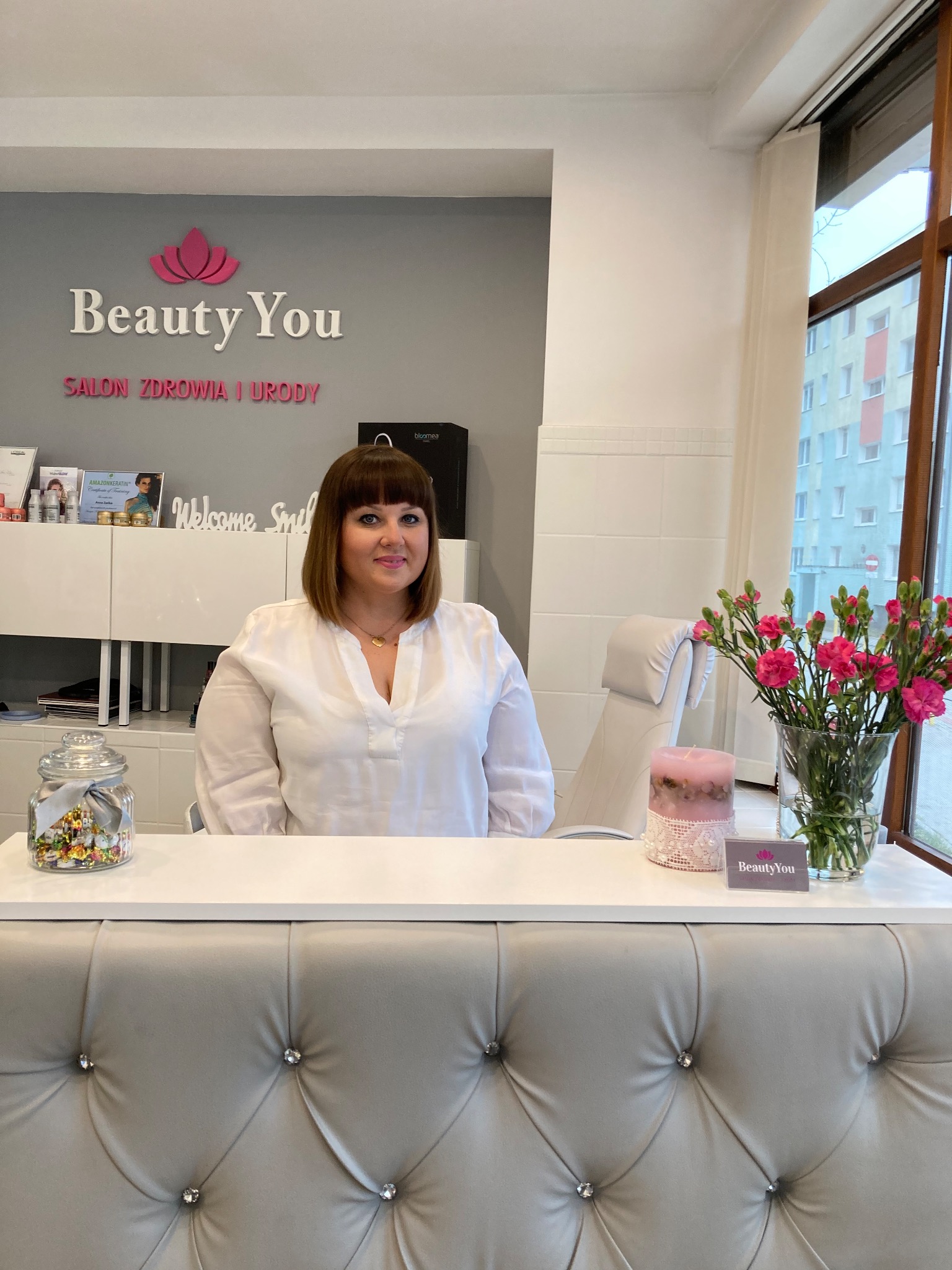 Recepcjonistka w salonie kosmetycznym Beauty You, za ladą z dekoracyjnym obiciem, w tle logo i wystrój wnętrza.