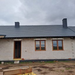 D.S DACH - Dom w trakcie budowy z ciemnoszarą dachówką i kominami z blachy, widoczne okna z drewnianą stolarką i ściany z betonu komórkowego.