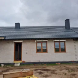 Dom w trakcie budowy z ciemnoszarą dachówką i kominami z blachy, widoczne okna z drewnianą stolarką i ściany z betonu komórkowego.