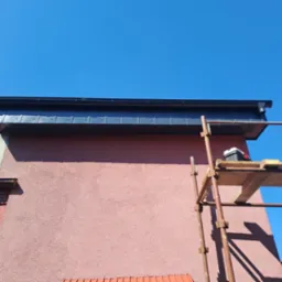 Zamontowany system orynnowania w kolorze grafitowym na tle różowej elewacji budynku, widoczne rusztowanie z narzędziami.