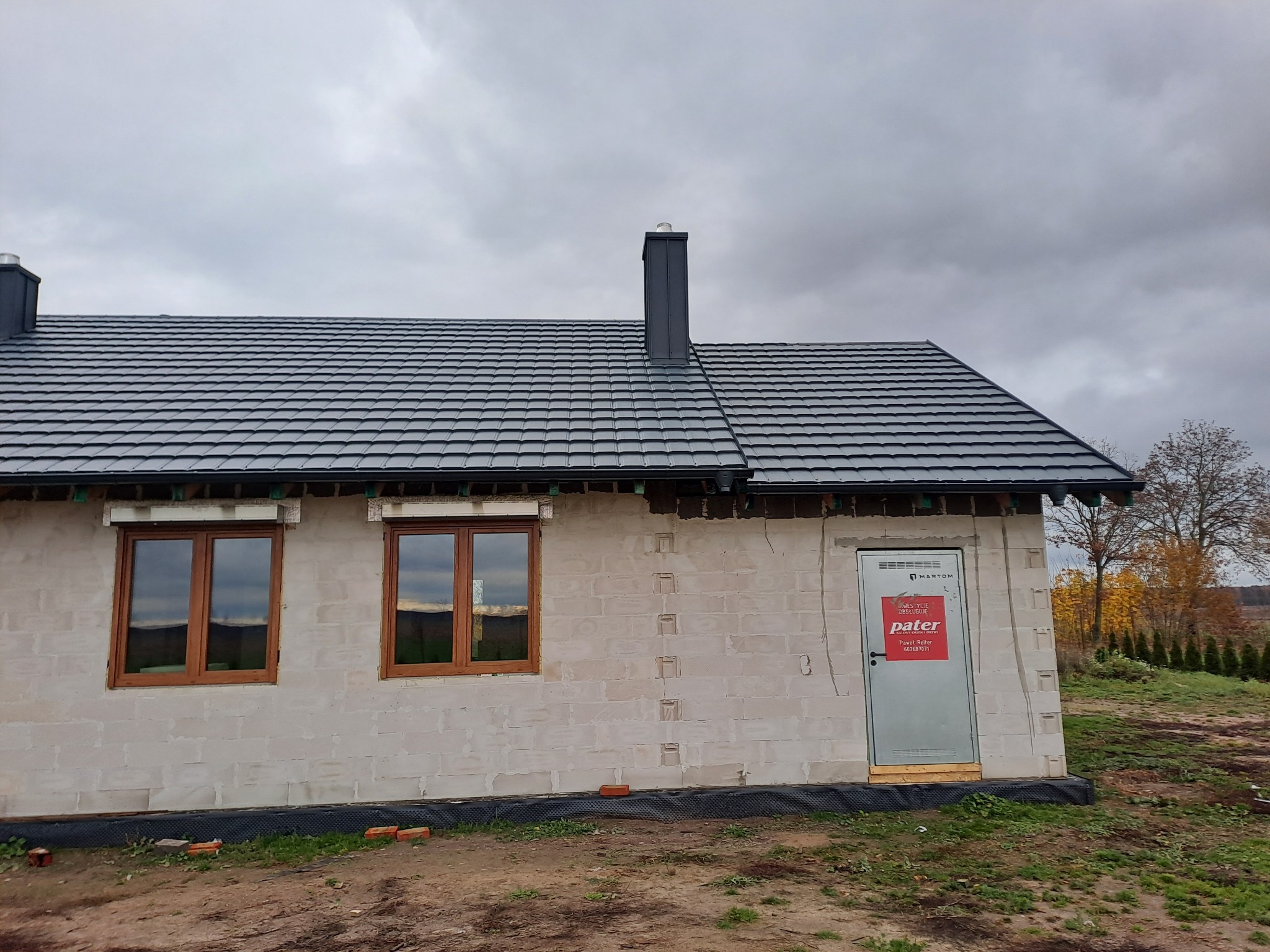 Dom w stanie surowym zamkniętym z nowym, ciemnoszarym dachem z blachodachówki modułowej i kominami w kolorze dachu, widoczne brązowe okna i tymczasowe drzwi wejściowe z naklejonym plakatem reklamowym.