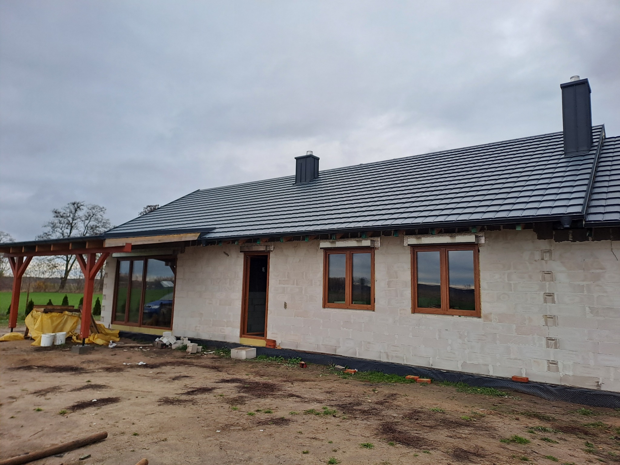 Dom jednorodzinny w trakcie budowy z nowym, grafitowym dachem z dachówki ceramicznej i kominem, widoczne okna z brązowymi ramami i częściowo zadaszony taras, teren wokół domu niezagospodarowany.