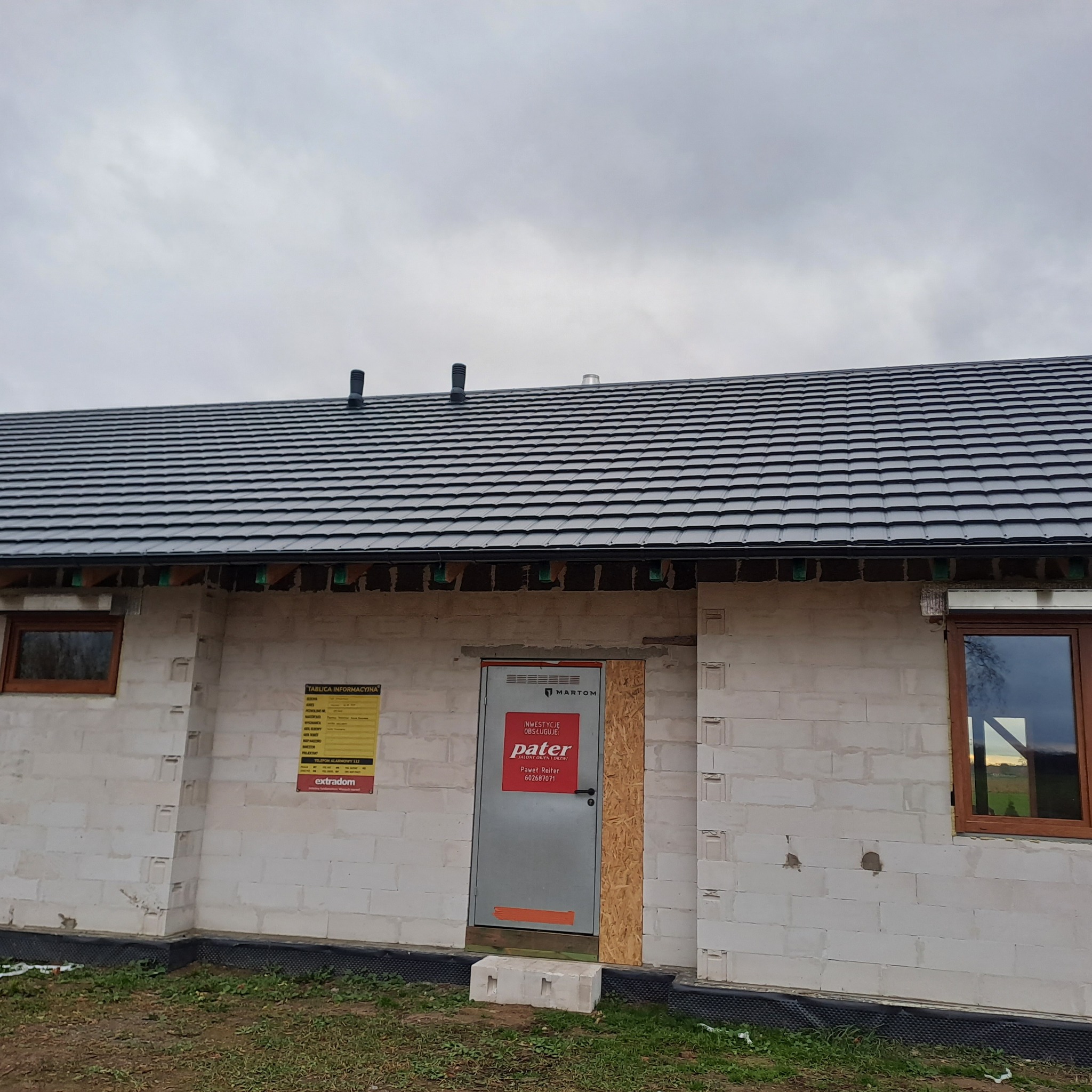 Nowy dach z dachówki ceramicznej na surowym budynku z białego bloczka, widoczne kominy wentylacyjne, okna z drewnianą ramą i tablica informacyjna budowy.