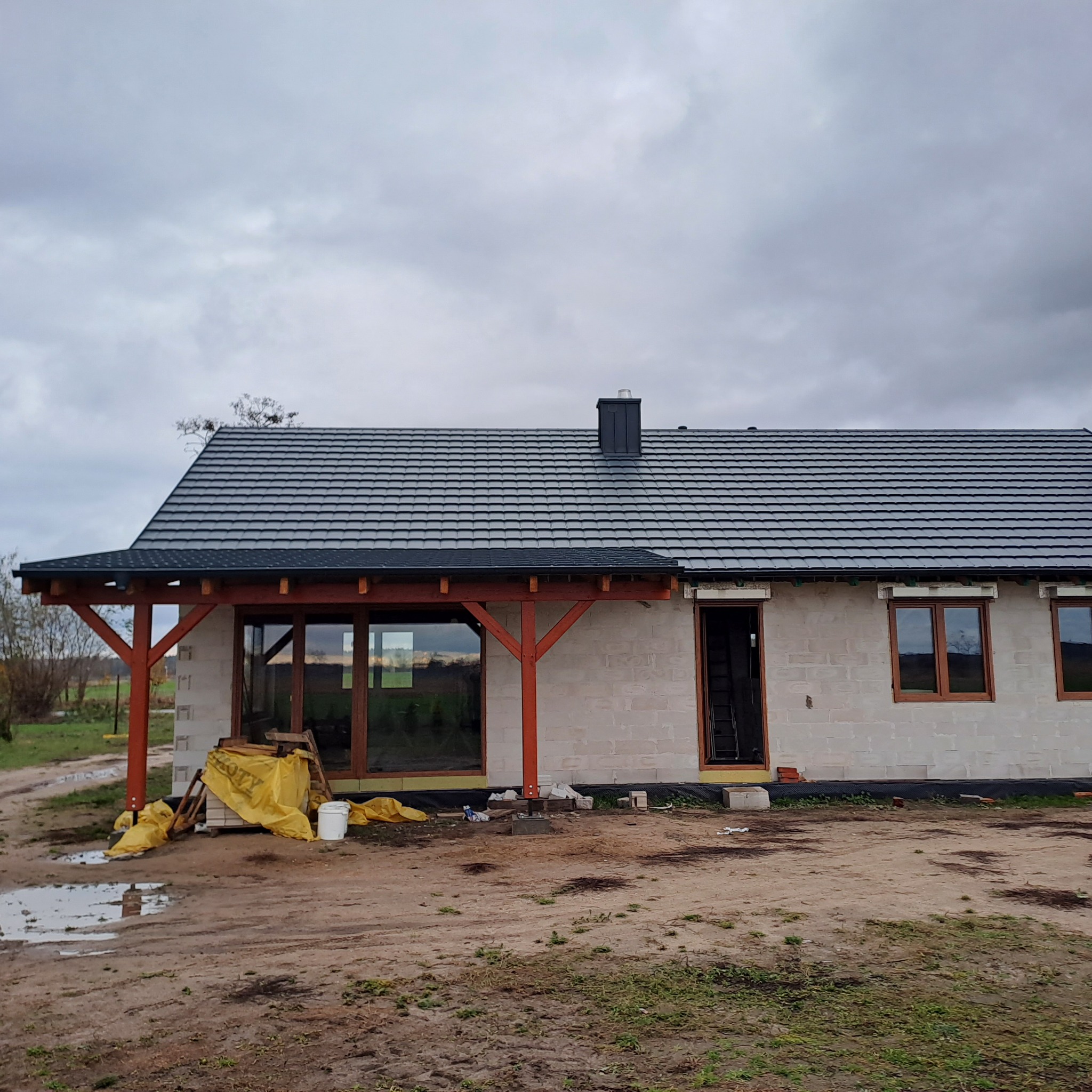 Dom jednorodzinny w trakcie budowy, widoczny szary dach z blachodachówki, surowy stan ścian z betonu komórkowego, drewniana konstrukcja zadaszenia tarasu, pochmurne niebo.