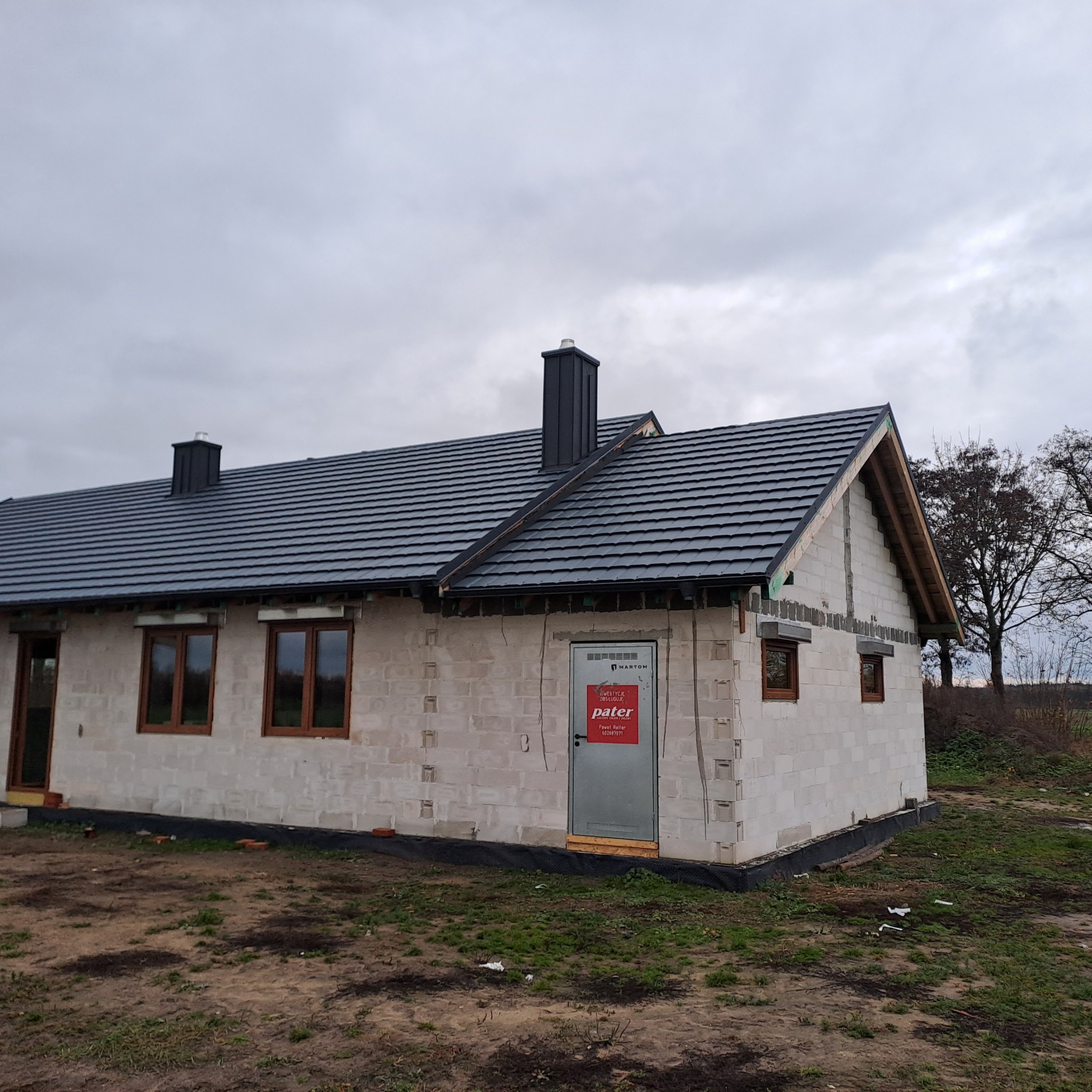 Dom jednorodzinny w trakcie budowy, widoczny nowy dach z ciemnej dachówki ceramicznej, komin z blachy, ściany z bloczków betonu komórkowego, okna z brązowymi ramami, drzwi z naklejoną reklamą.
