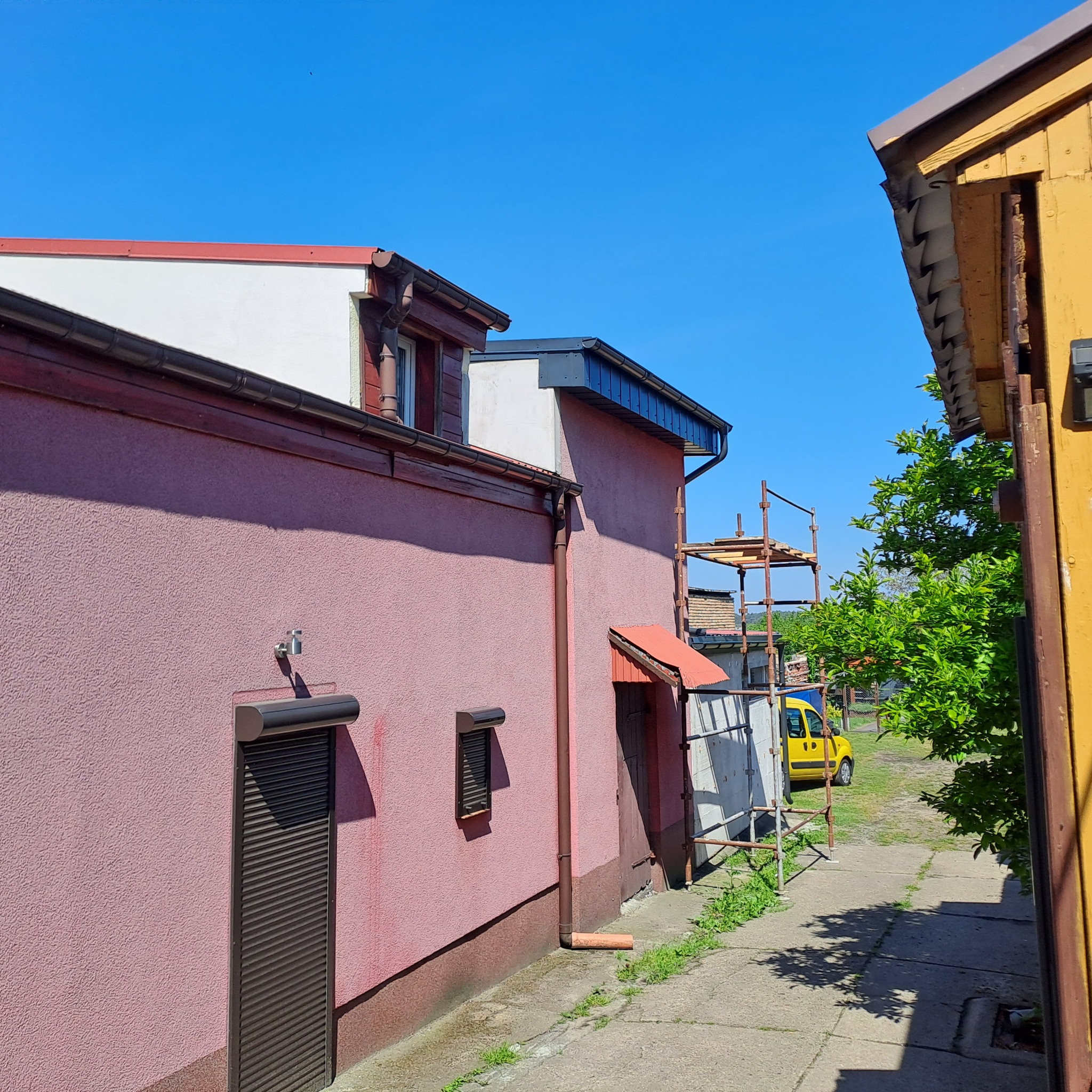 Fragment elewacji budynku z rynną, roletą okienną i fragmentem dachu oraz rusztowaniem w tle, widziany w słoneczny dzień.