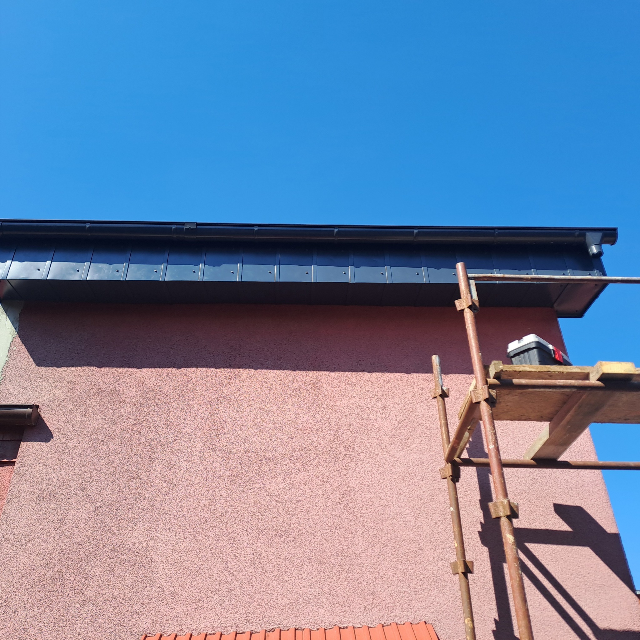 Zamontowany system orynnowania w kolorze grafitowym na tle różowej elewacji budynku, widoczne rusztowanie z narzędziami.