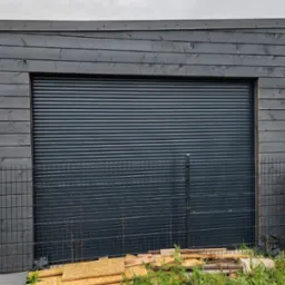 Zamknięta, szara brama garażowa rolowana, zamontowana w budynku z ciemnego drewna, z widocznym fragmentem metalowego ogrodzenia i stertą drewnianych odpadów na trawie.