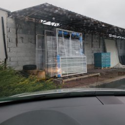 VINSTA SPÓŁKA CYWILNA WOJCIECH PAJĄK, DMYTRO ZHULINSKYI - Zabezpieczone folią ramy okienne z niebieskimi profilami, składowane pod zadaszeniem magazynu budowlanego w pochmurny dzień, widok zza szyby samochodu.