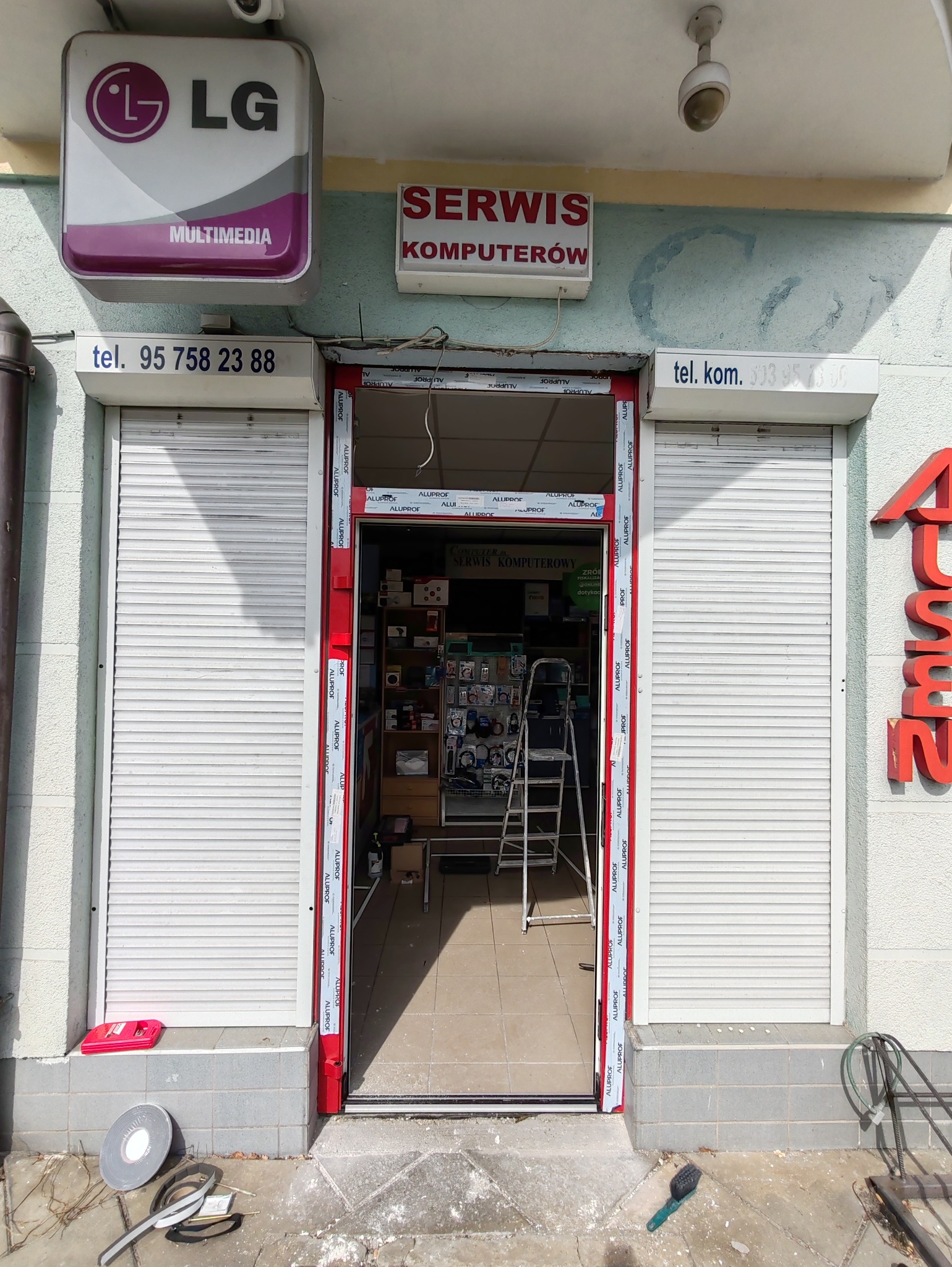 Montaż aluminiowej ramy drzwiowej wejścia do serwisu komputerowego z częściowo opuszczonymi roletami zewnętrznymi, widoczne narzędzia i drabina wewnątrz.