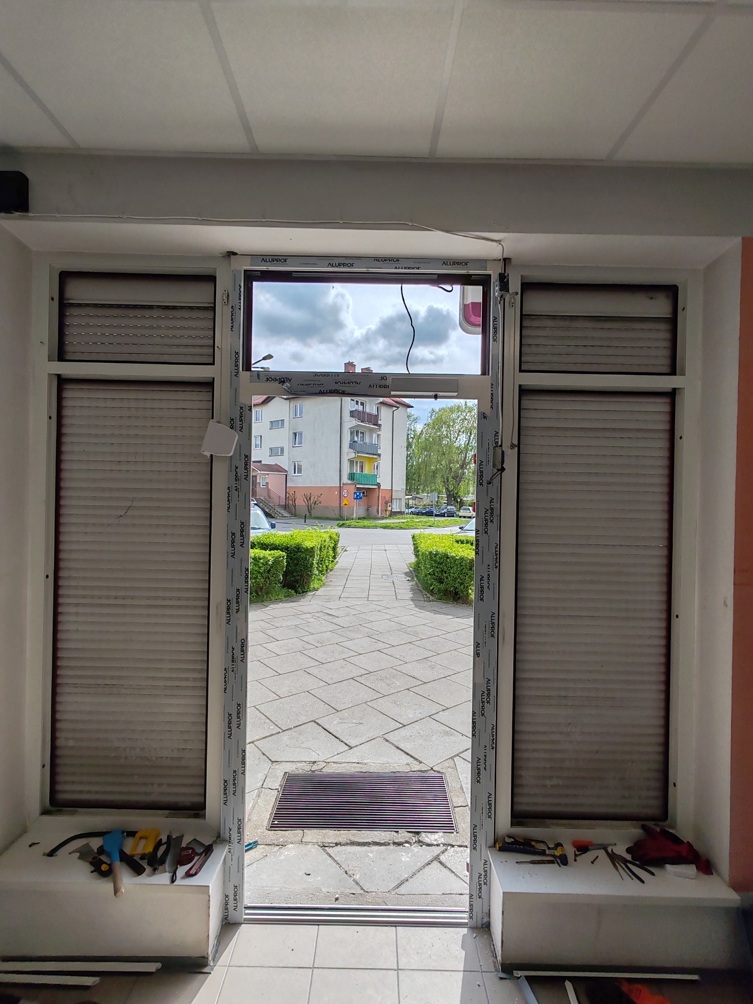 Montaż ramy okiennej z roletami zewnętrznymi w lokalu, widok na chodnik i budynki mieszkalne, narzędzia na parapecie.