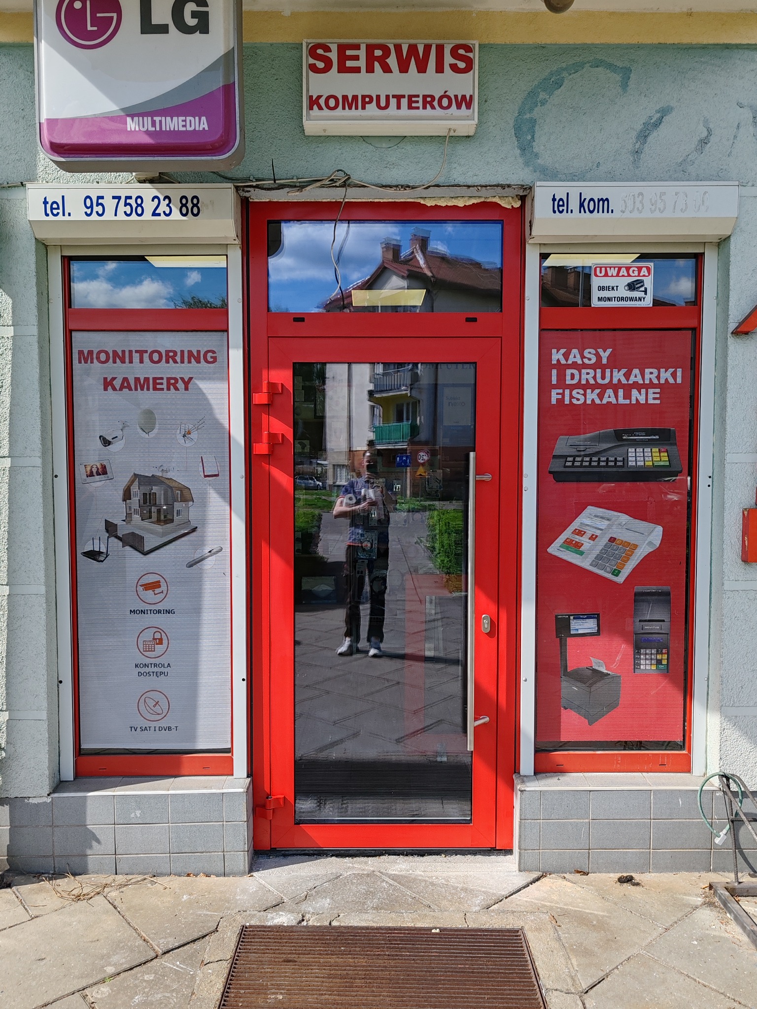 Czerwone drzwi wejściowe do serwisu komputerowego z reklamami monitoringu kamer i kas fiskalnych na szybach, odbicie osoby w drzwiach.