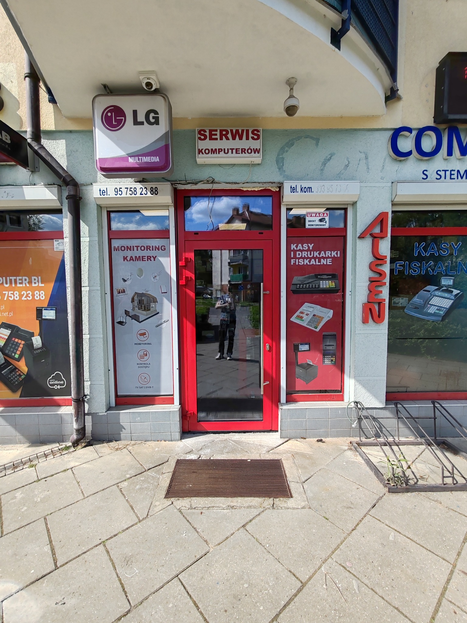Fasada lokalu usługowego z czerwonymi ramami okiennymi i drzwiowymi, oferującego serwis komputerowy, monitoring kamer oraz kasy i drukarki fiskalne, widoczny szyld LG Multimedia i kamery...