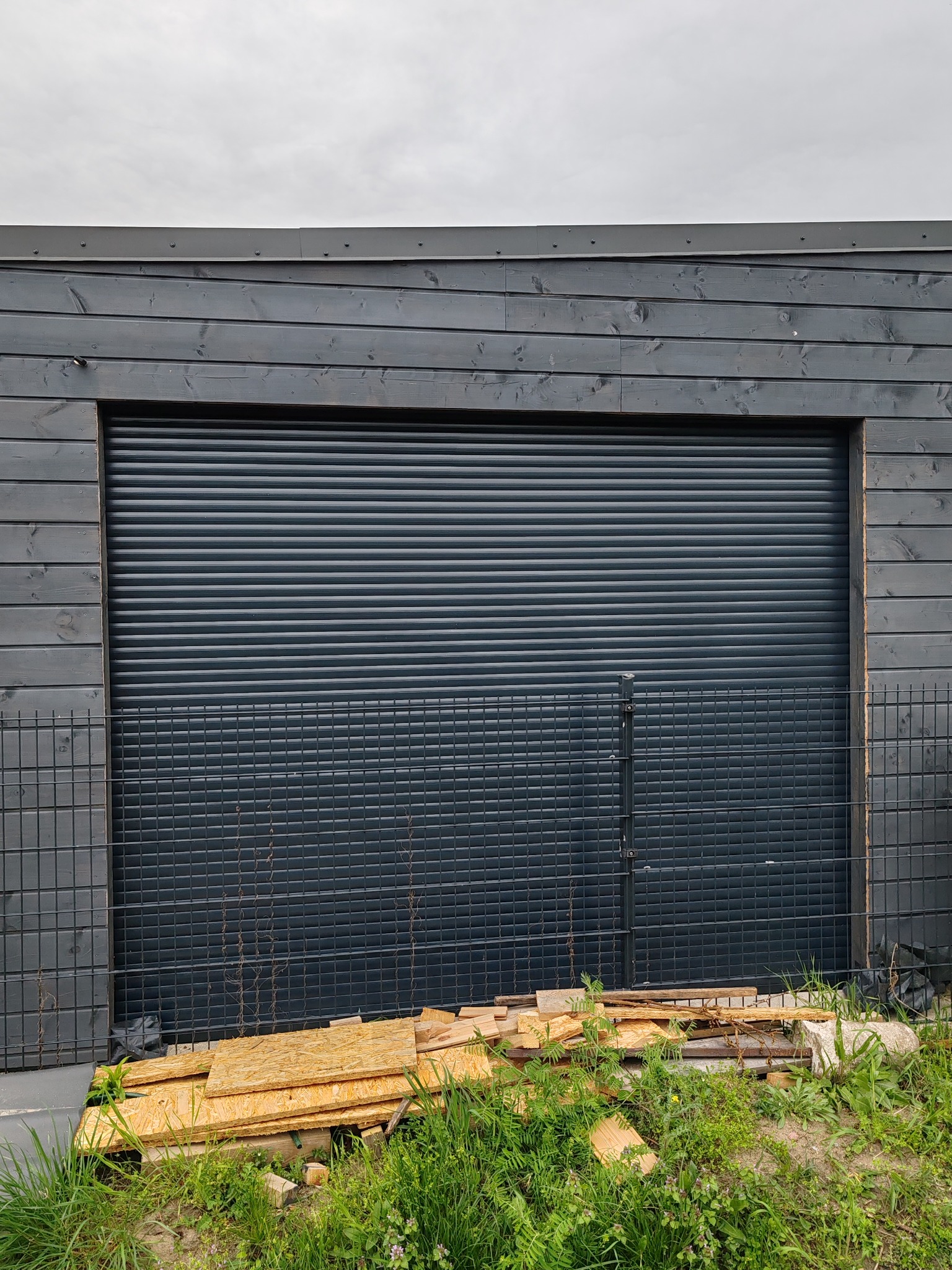 Zamknięta, szara brama garażowa rolowana, zamontowana w budynku z ciemnego drewna, z widocznym fragmentem metalowego ogrodzenia i stertą drewnianych odpadów na trawie.