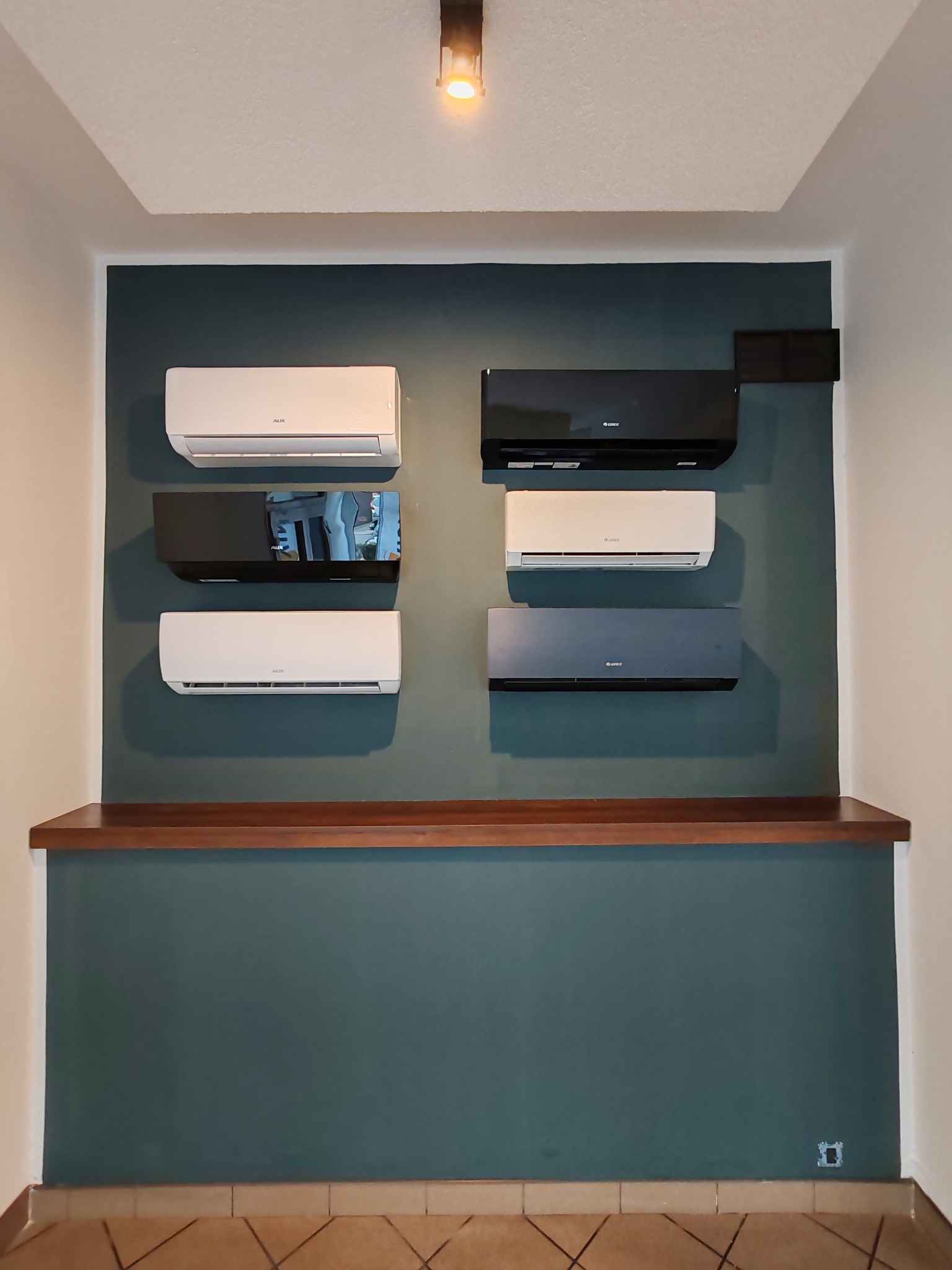 Ekspozycja klimatyzatorów różnych marek i kolorów (biały, czarny, szary) zamontowanych na ścianie w pomieszczeniu, pod drewnianą półką.