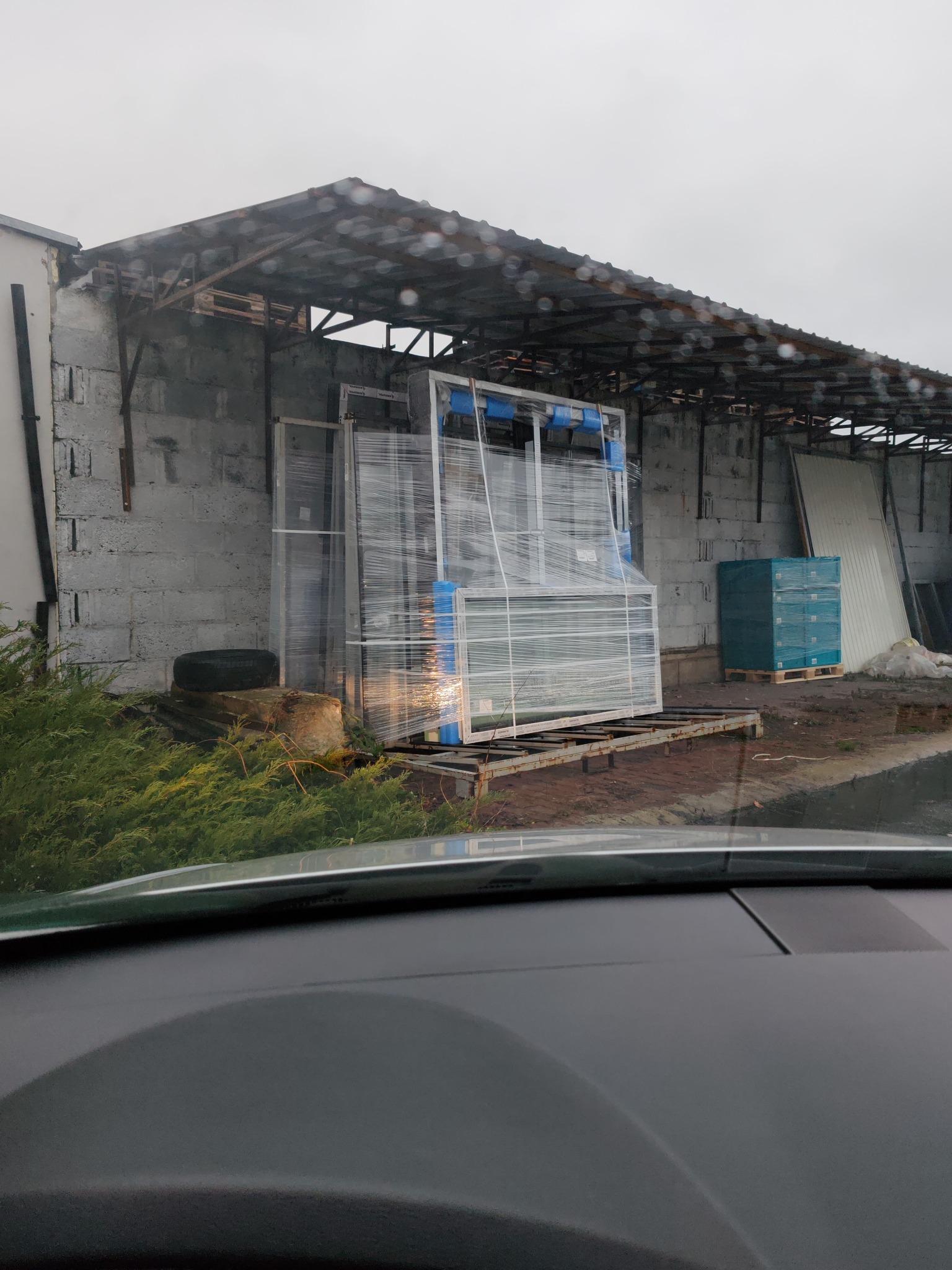 Zabezpieczone folią ramy okienne z niebieskimi profilami, składowane pod zadaszeniem magazynu budowlanego w pochmurny dzień, widok zza szyby samochodu.