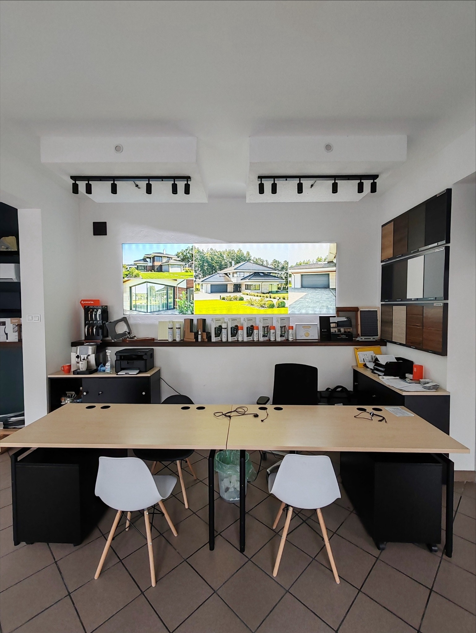 Wnętrze biura z ekspozycją domów jednorodzinnych na ekranie, próbki wykończeń na ścianie, biurko z krzesłami i akcesoriami biurowymi.