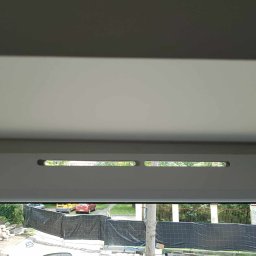 Darek Godek - Widok z okna po naprawie, z widocznymi dwoma otworami wentylacyjnymi w górnej ramie okna, na zewnątrz widoczny ogród i zaparkowane samochody za czarną siatką.