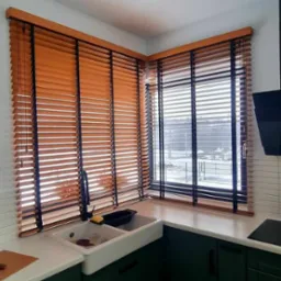 Drewniane żaluzje poziome w kolorze miodowym z czarnymi sznurkami, zamontowane w narożnym oknie kuchennym nad zlewem farmerskim i zielonymi szafkami.