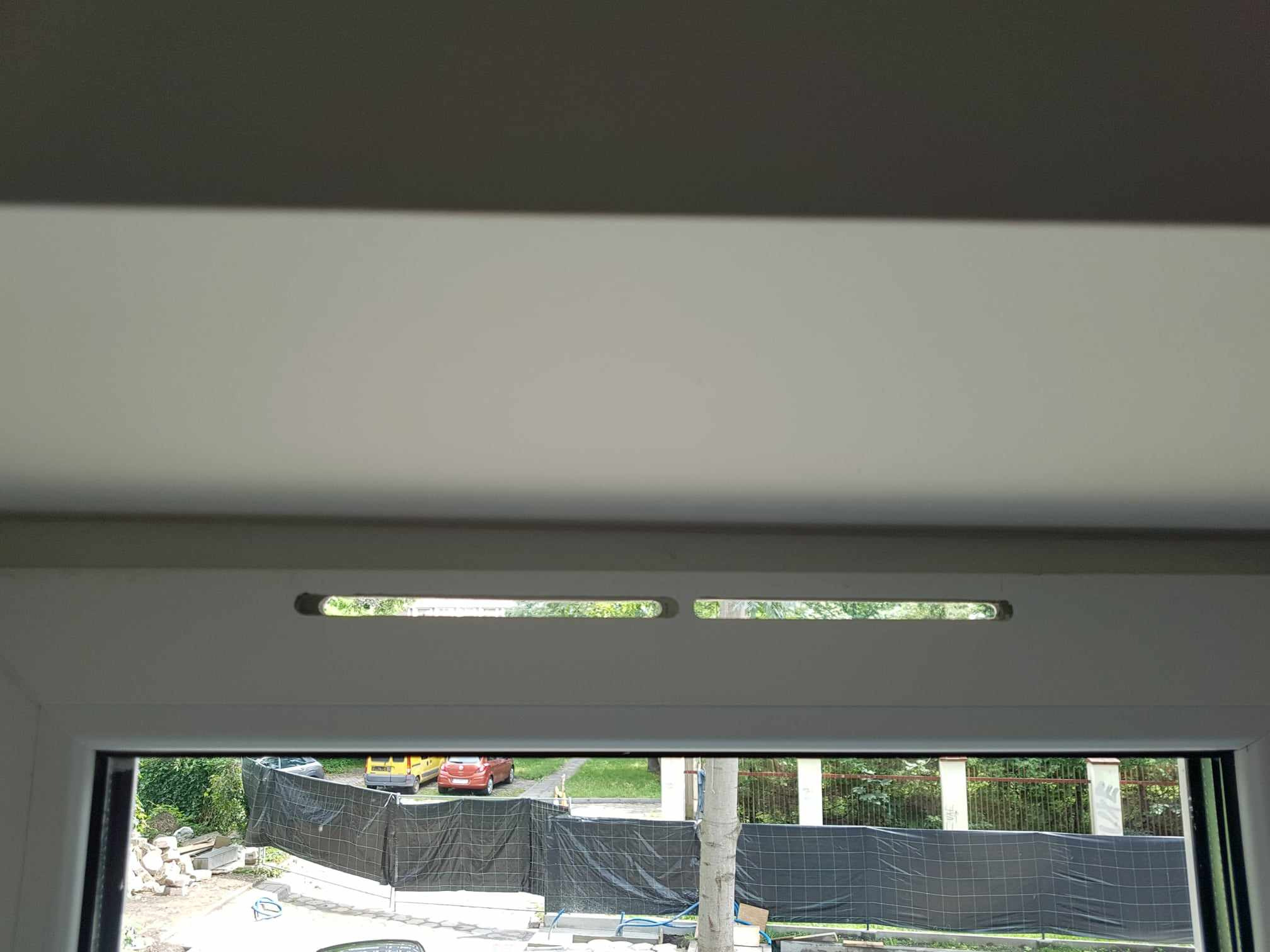 Widok z okna po naprawie, z widocznymi dwoma otworami wentylacyjnymi w górnej ramie okna, na zewnątrz widoczny ogród i zaparkowane samochody za czarną siatką.