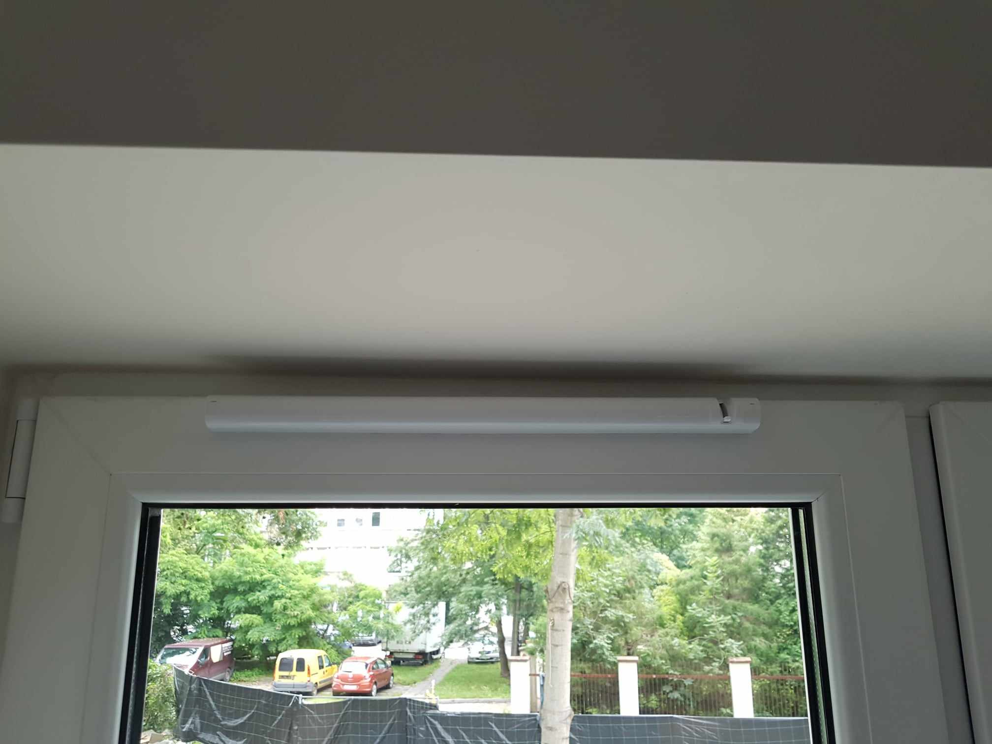 Widok z okna z zamontowaną roletą zwiniętą u góry, za oknem zieleń drzew i zaparkowane samochody.