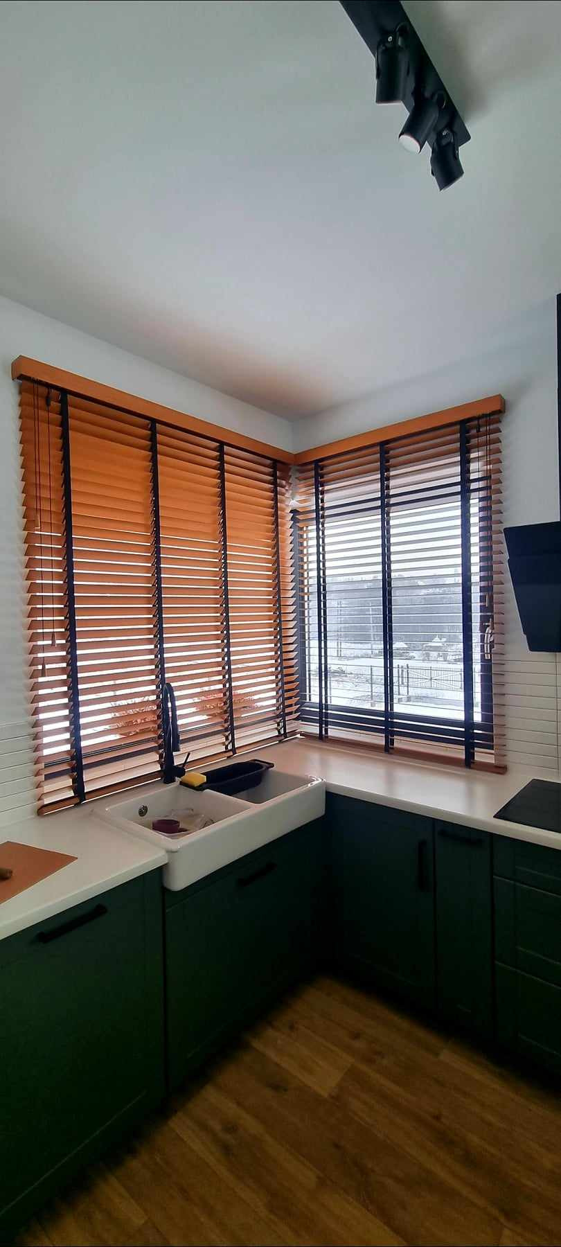 Drewniane żaluzje poziome w kolorze miodowym z czarnymi sznurkami, zamontowane w narożnym oknie kuchennym nad zlewem farmerskim i zielonymi szafkami.