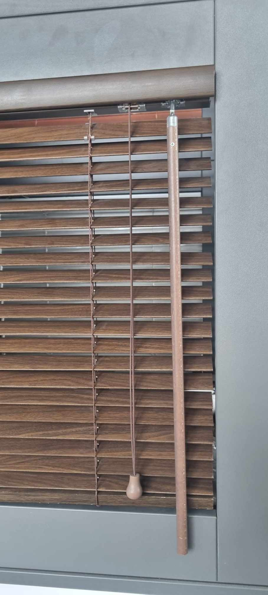 Drewniane żaluzje poziome w kolorze orzecha włoskiego z szarym obramowaniem okna, widoczne sznurki i drewniany uchwyt do regulacji.