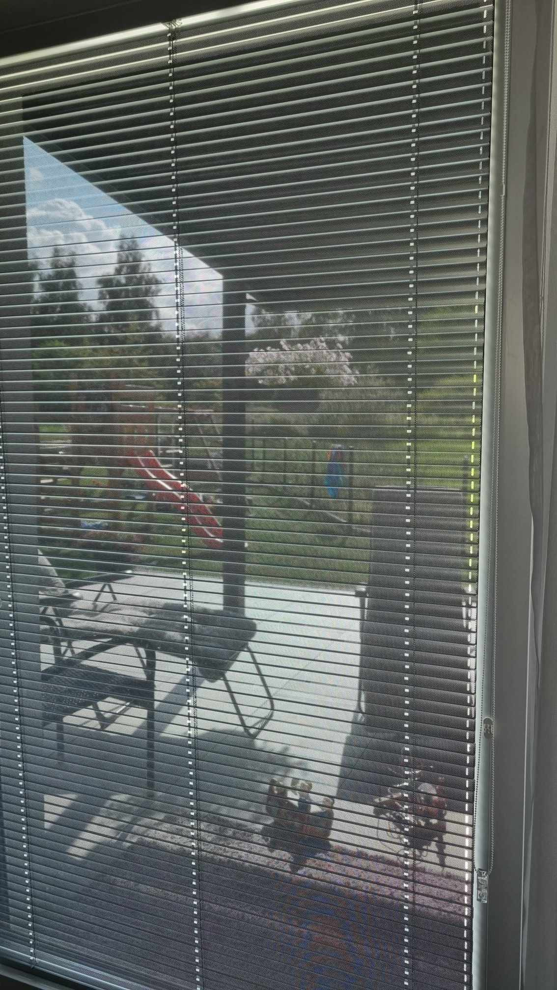 Zasłonięte okno z widokiem na taras i plac zabaw przez metalowe żaluzje, częściowo opuszczone.