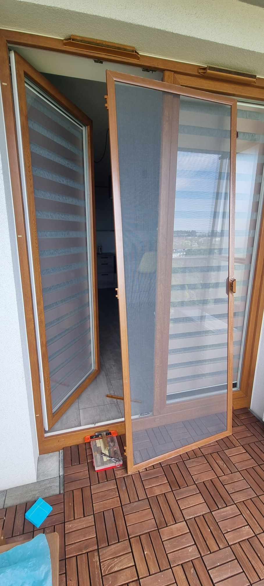 Brązowa rama moskitiery na tle otwartego okna z roletami dzień-noc i narzędziownika na drewnianej podłodze balkonu.