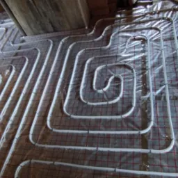 Białe rury ogrzewania podłogowego ułożone spiralnie na folii z siatką w nowym budownictwie, widoczne elementy konstrukcyjne budynku.